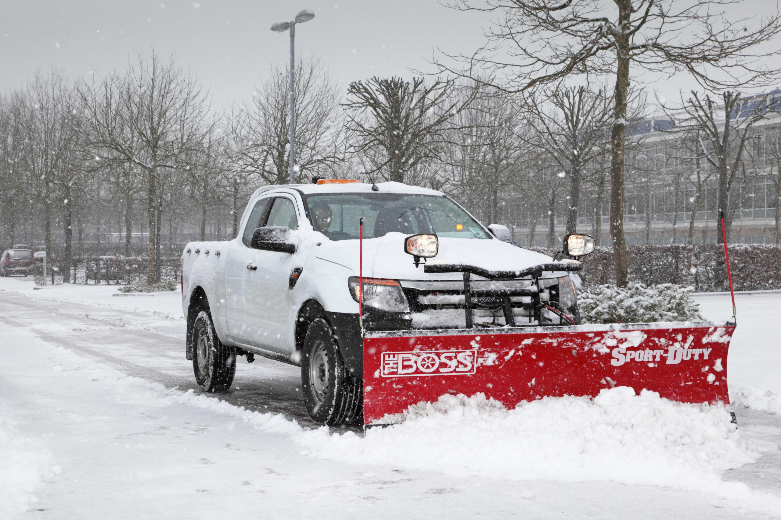 A white utility truck equipped with a red snow plough is clearing snow from a road during a heavy snowfall. The plough is pushing the snow to the side, creating a clear path. Snow is accumulating on the truck's bonnet, roof, and around the wheels, indicating continuous snowfall. In the background, bare trees and buildings are visible, partially obscured by the falling snow. The scene reflects a typical winter snow-clearing operation in a car park or urban area.